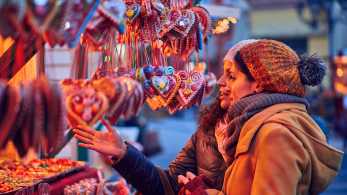 Two women admire a display of decorations at a Christmas market, surrounded by holiday cheer.