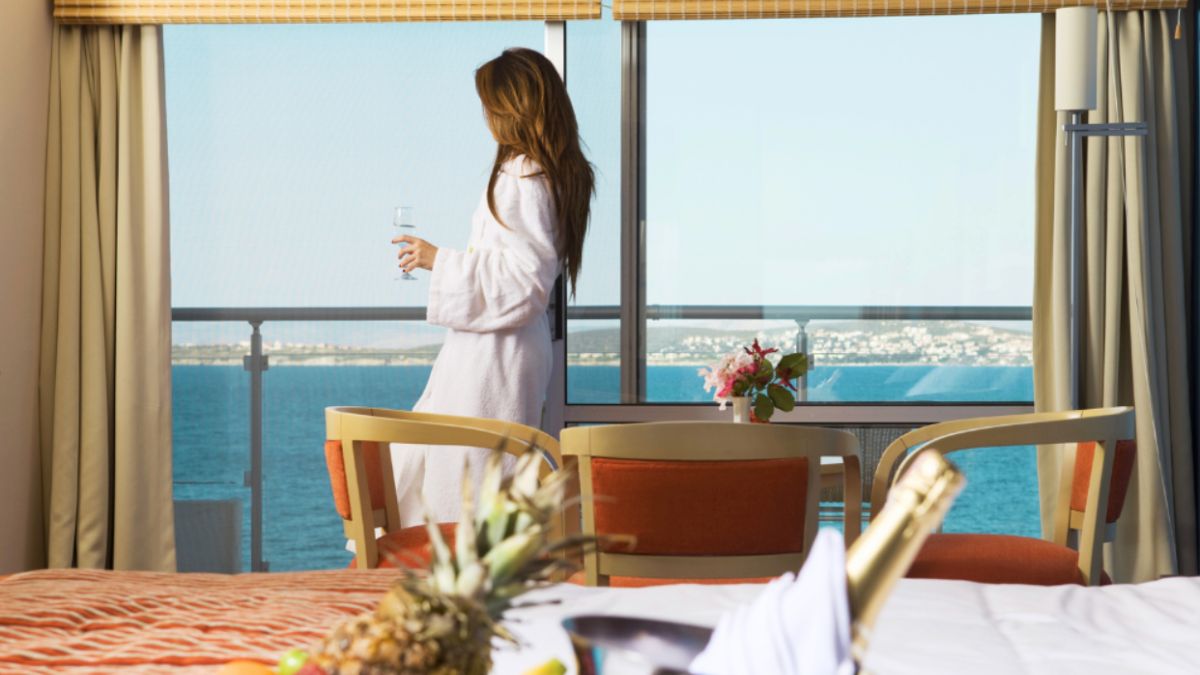 A woman in a robe stands by a window, looking at the ocean, representing the allure of luxury travel planning.