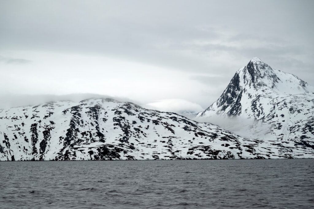 Snow-covered mountains rise from a dark, choppy sea near Greenland