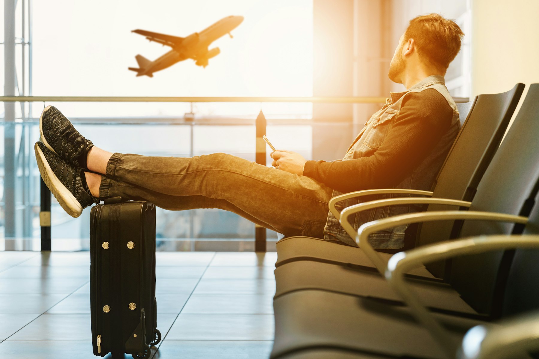 A traveller reclining across gate seating with his feet on a suitcase, watching an aircraft climb away, the free alternative to an airport lounge.