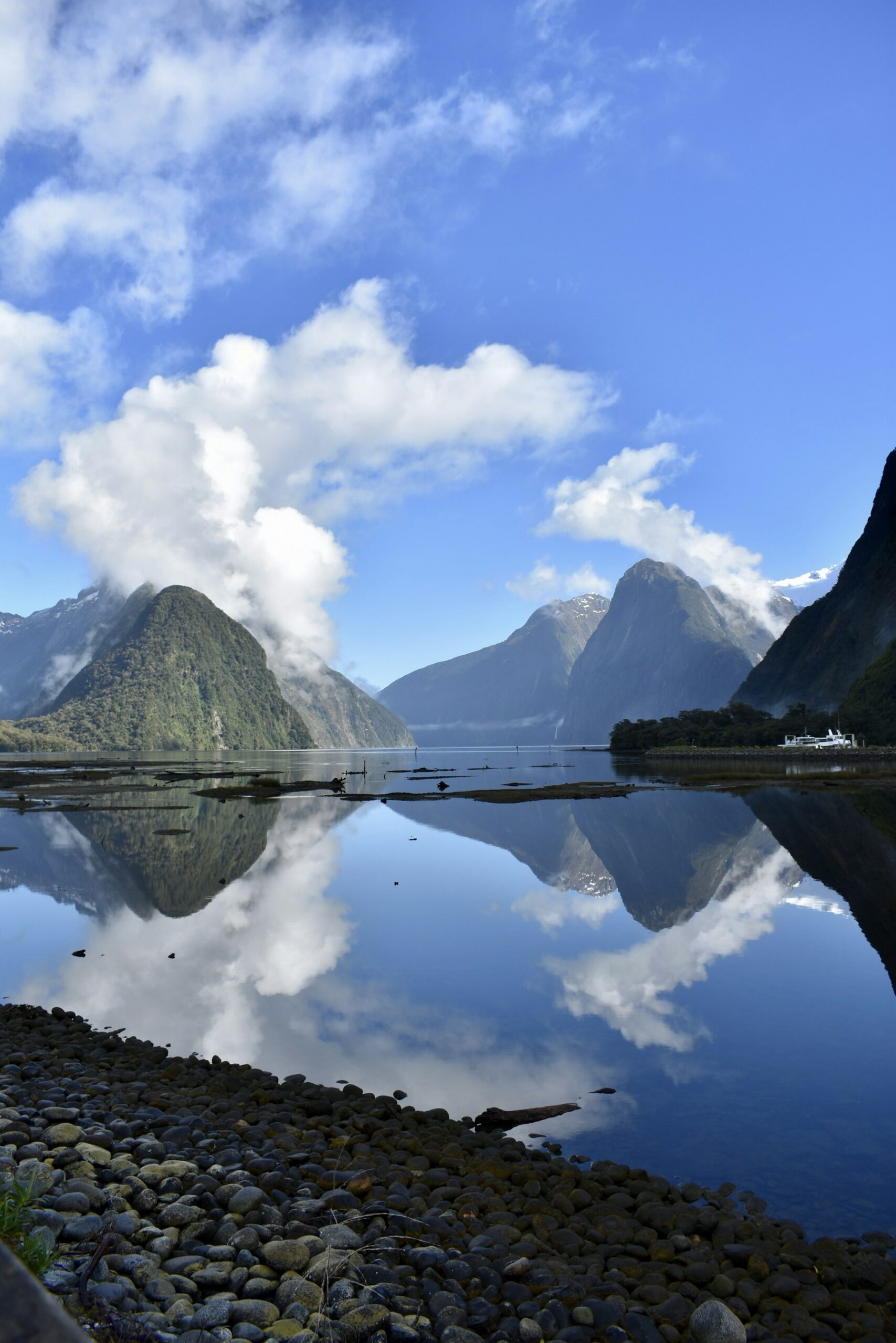 Milford Sound, Piopiotahi, at dead calm, with cloud spilling over the fiord walls and reflected in the water and a small vessel at the shoreline.