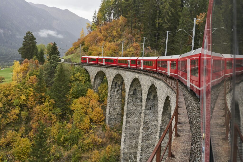Switzerland by train: a red Rhaetian Railway train crossing a tall stone-arched viaduct on the Albula line above an autumn forest near Bergun.