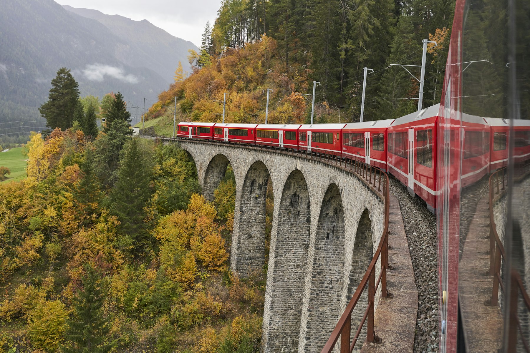 Switzerland by train: a red Rhaetian Railway train crossing a tall stone-arched viaduct on the Albula line above an autumn forest near Bergun.