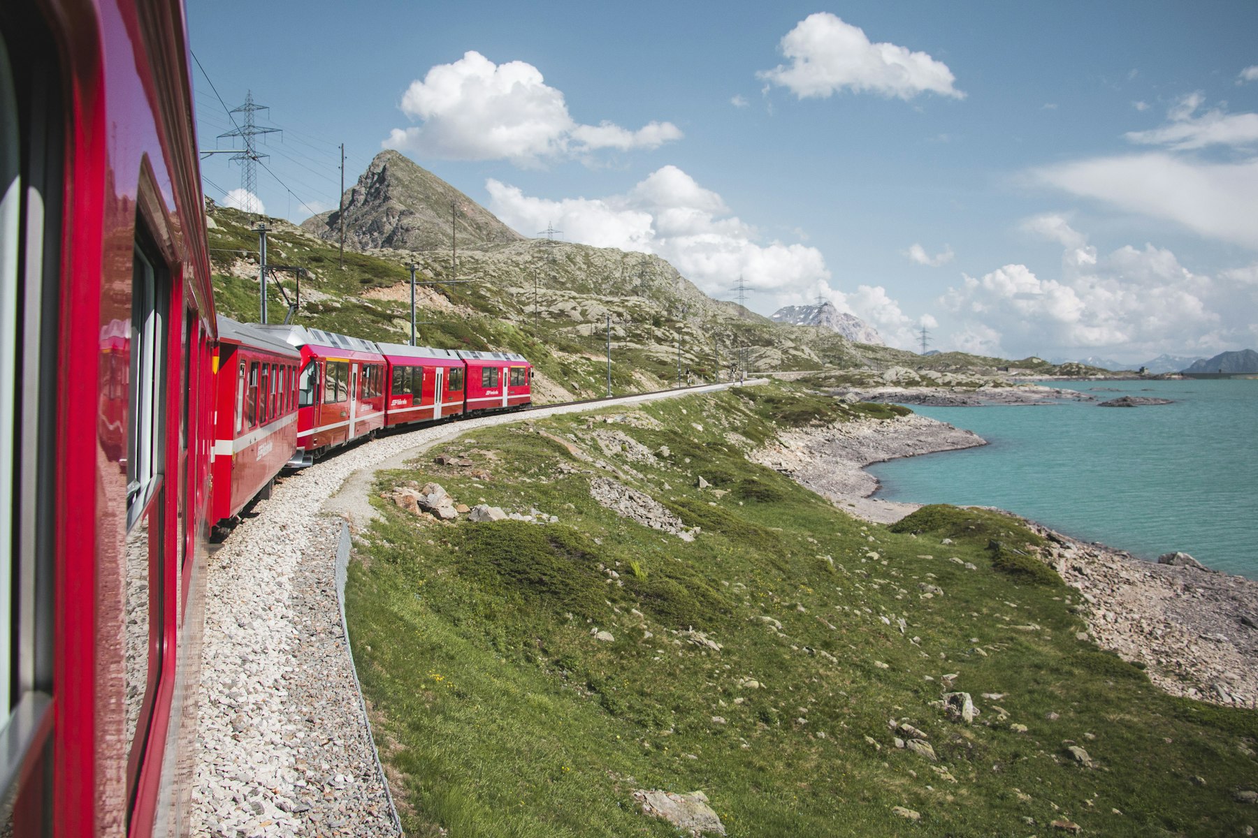 A red train curving along the shore of a turquoise glacial lake on the Bernina Pass, photographed from an open carriage window.