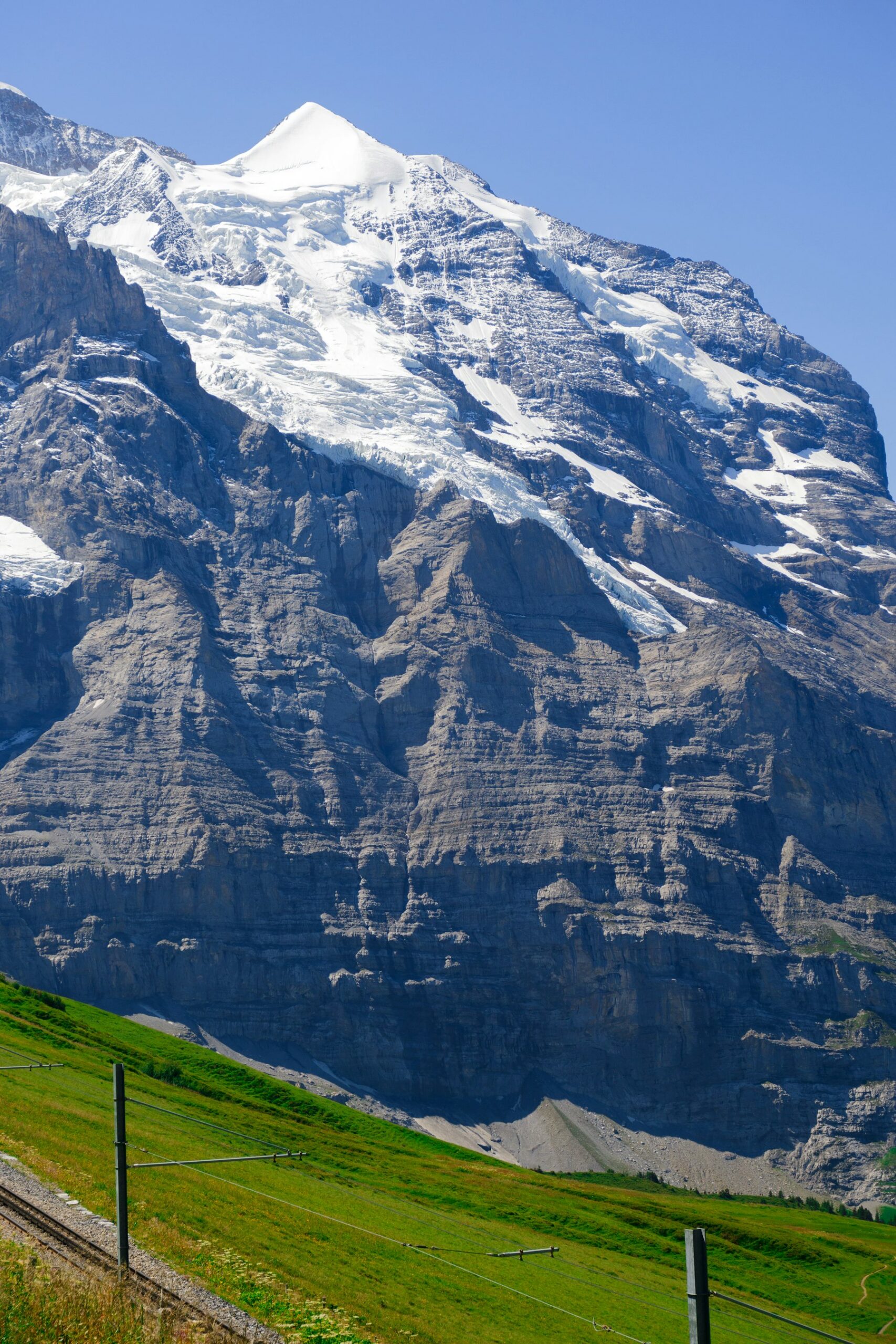 A glaciated mountain wall above a green alpine meadow, with a railway line and catenary poles running along the slope below it at Lauterbrunnen.